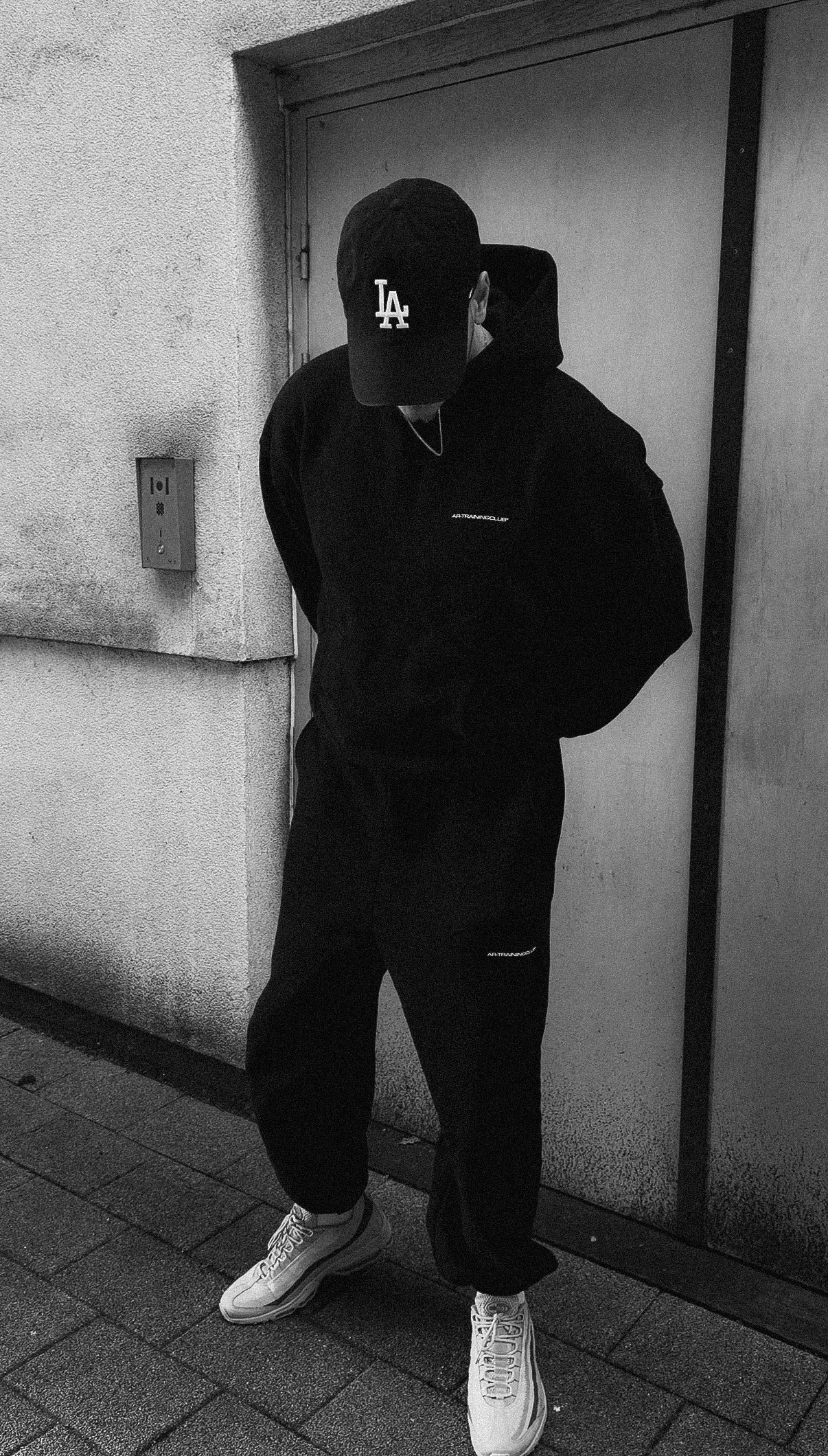 Person wearing a black hoodie and cap with 'LA' logo, standing against a wall.
