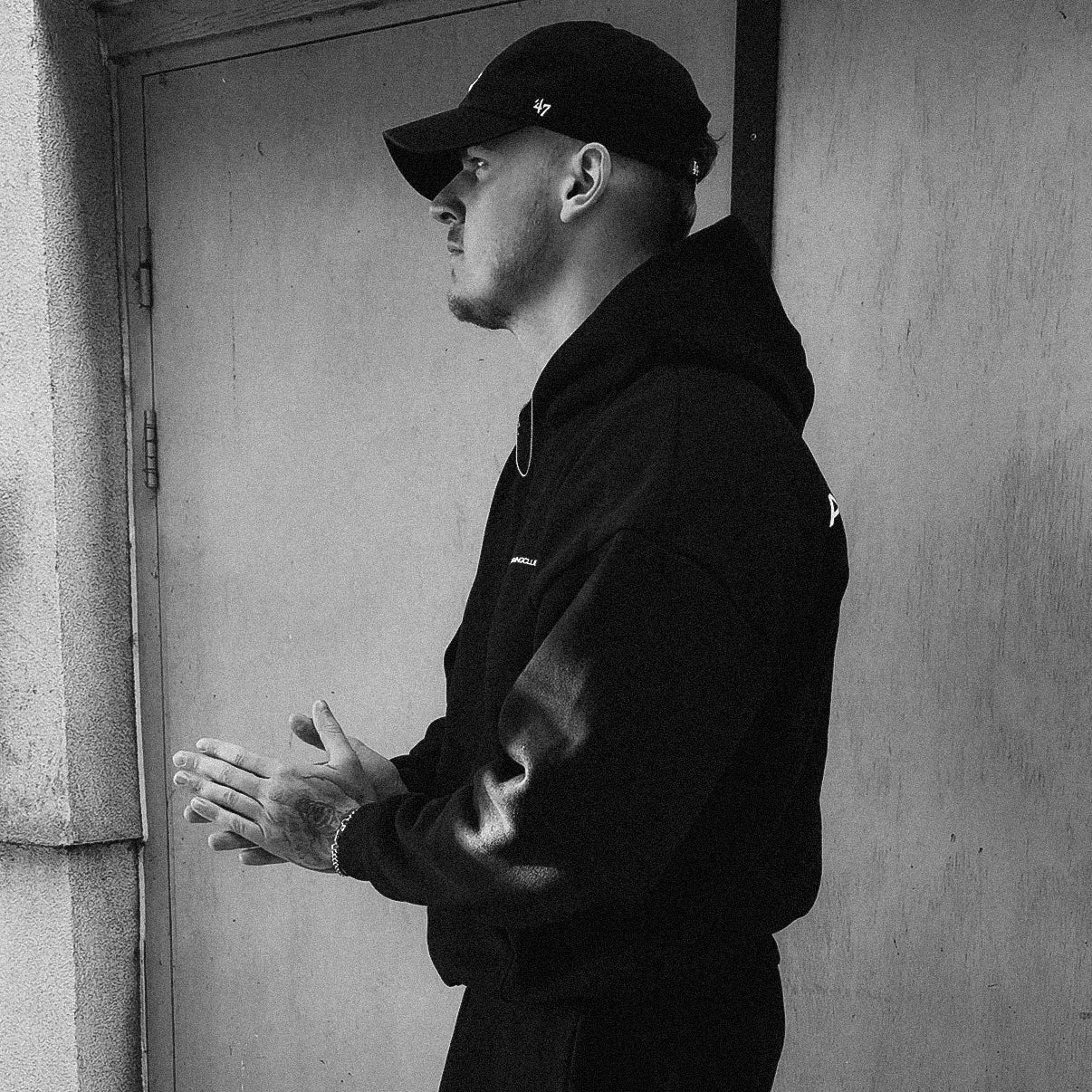 Man in black outfit standing against a plain wall