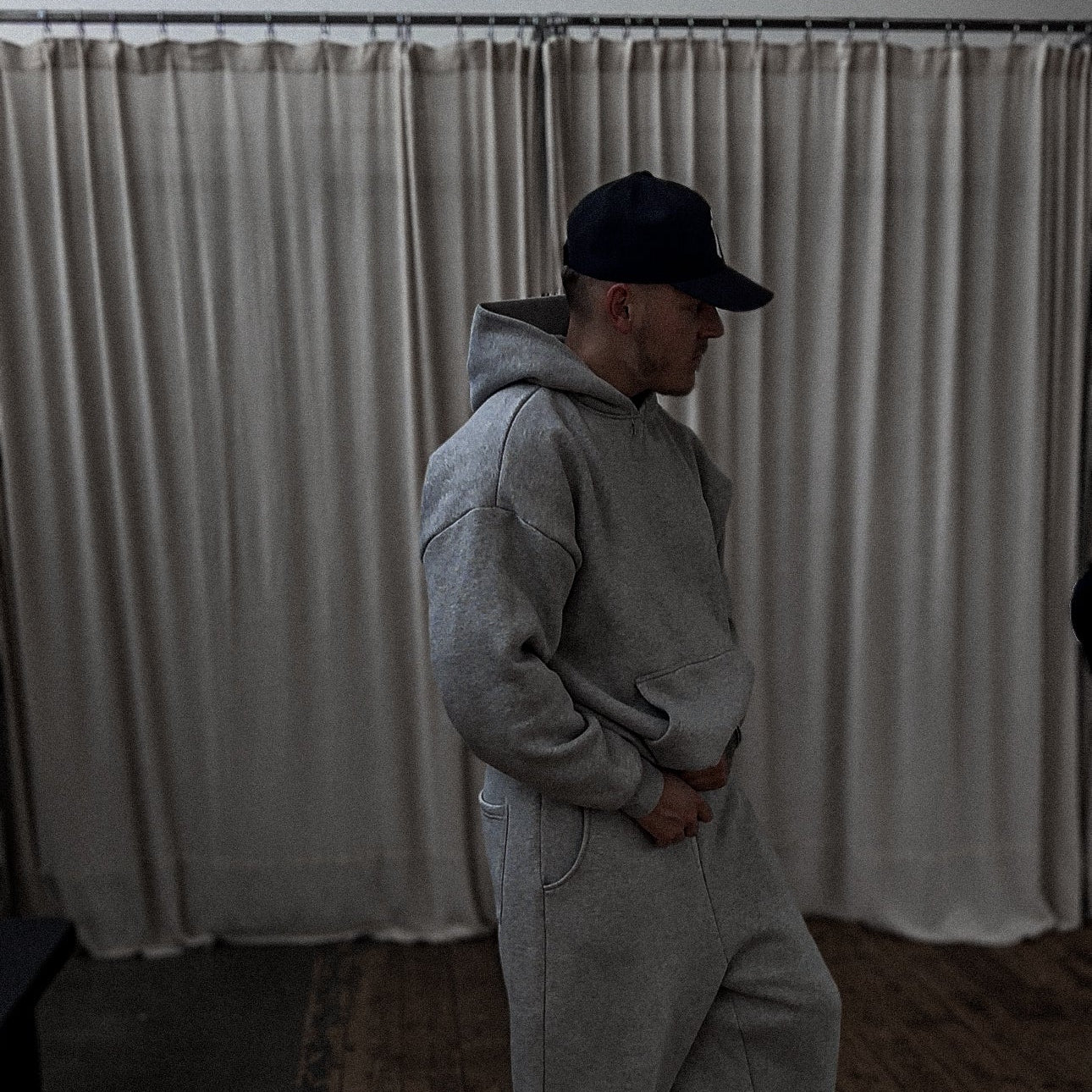 Person wearing a gray hoodie and pants with a black cap in a room with white curtains.